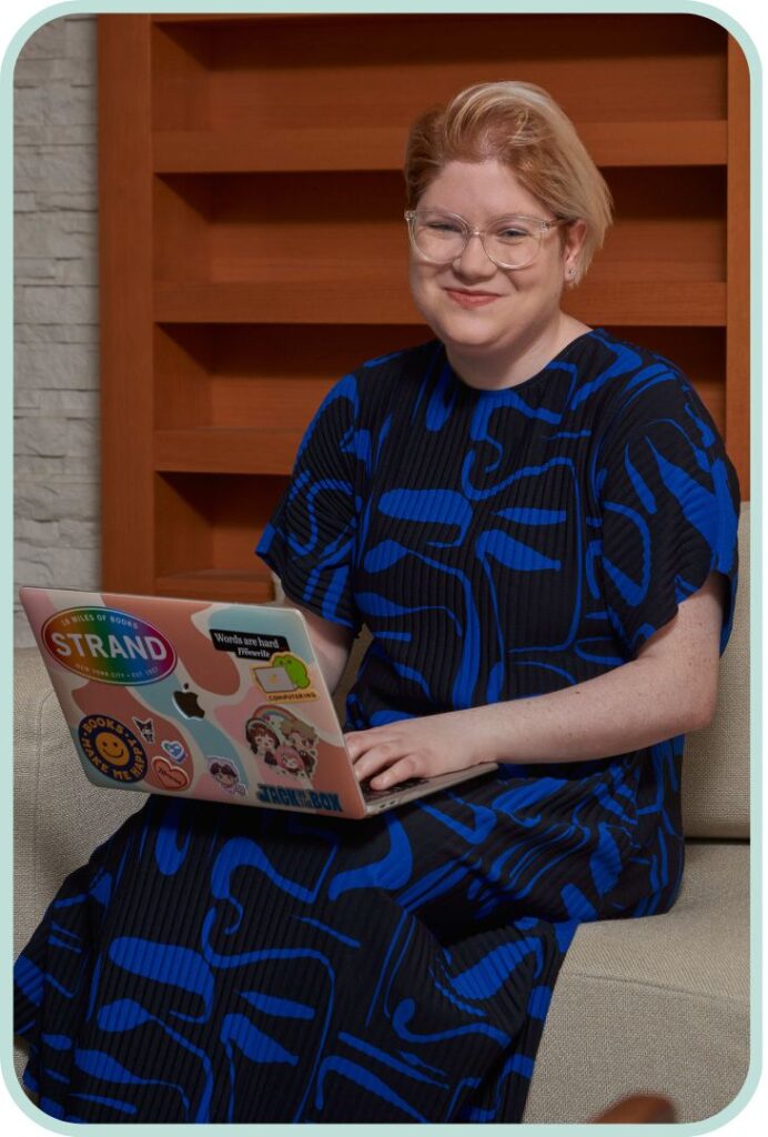 Image of Shelly Zev smiling with a laptop full of colourful stickers. They are wearing a blue and black patterened dress and have short light hair. Shelly is a Toronto-based book editor extraordinaire! They have extensive experience as a romance novel editor, poetry editor, fiction editor, and more! 
